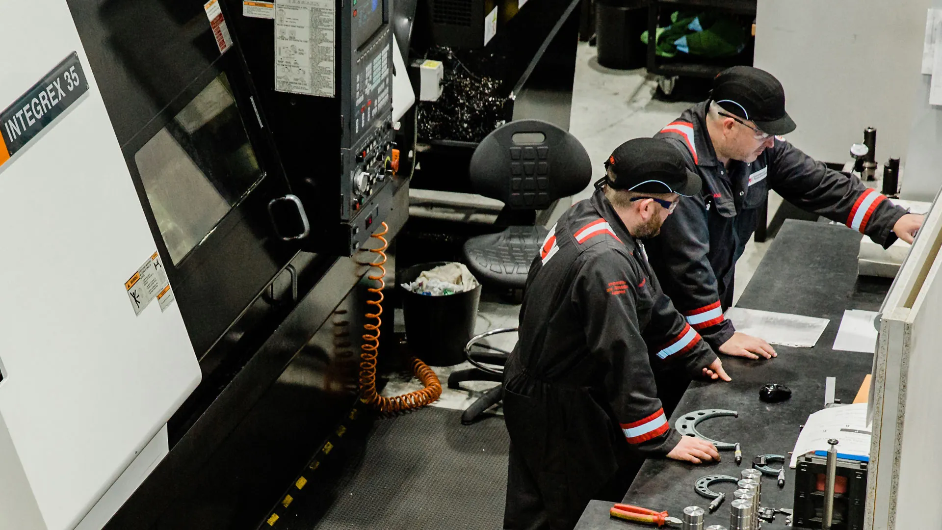 Two machinists in shop coveralls at an inspection bench beside a CNC lathe, checking finished parts against gauges.