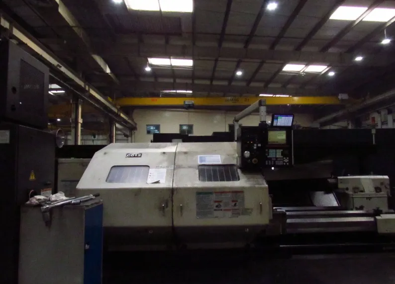 An older CNC lathe on a shop floor, its original control panel still in service under an overhead crane.