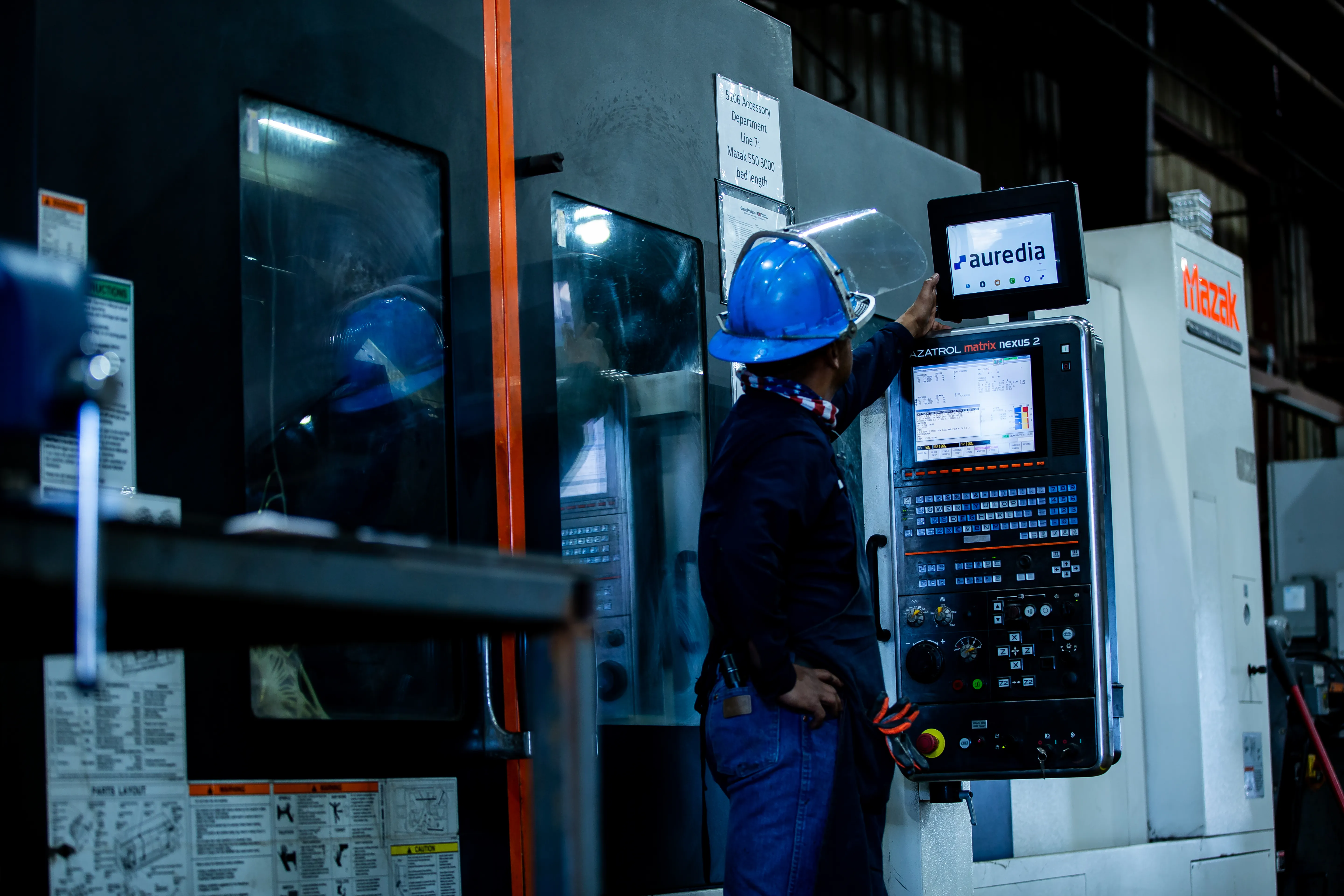 A machinist in a blue hard hat reaching up to a tablet mounted on a Mazak machining centre, the Auredia interface open on the screen.