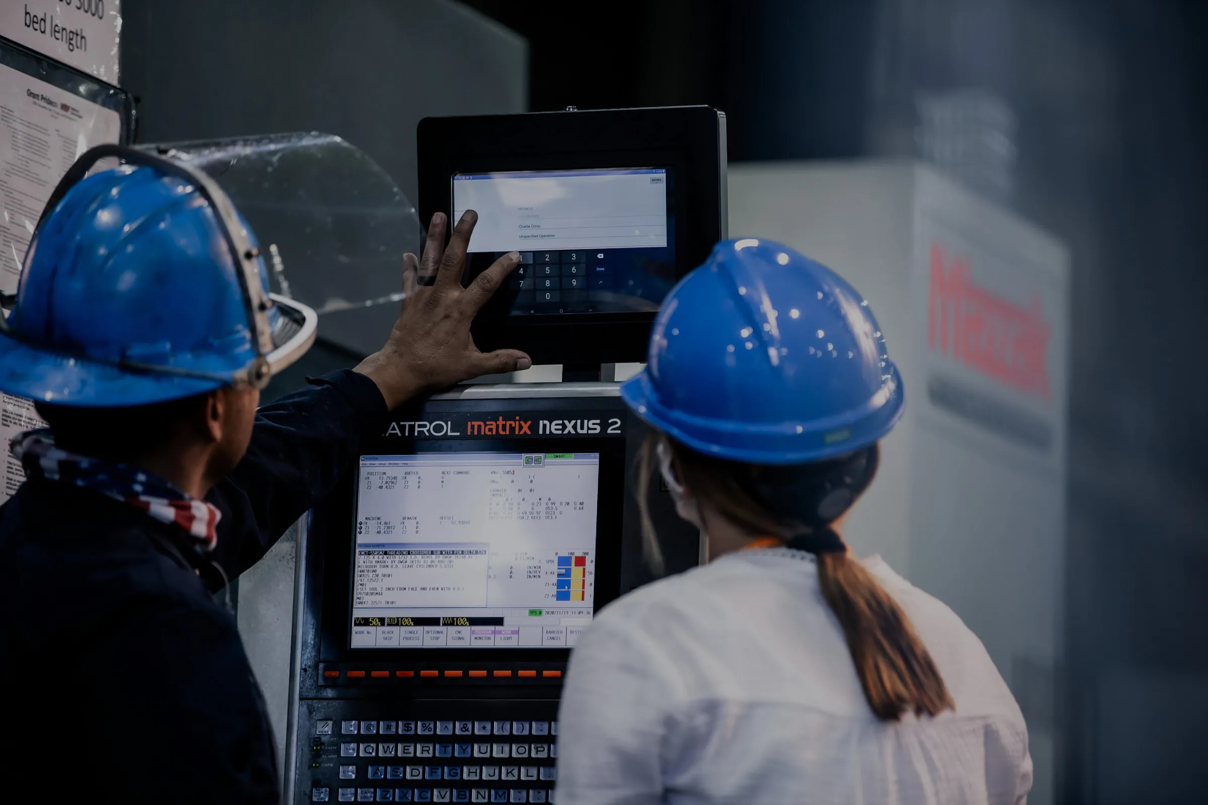 Two machinists in blue hard hats at a CNC control station, one reaching up to a touchscreen showing an Auredia work order while the machine's own display runs beneath it.